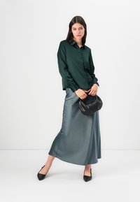 Blusa di raso verde con chiusura a bottoni, abbinata a una gonna maxi di raso argento. La modella tiene una borsa nera, indossando scarpe nere a punta.