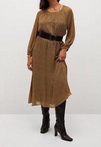 Brązowa sukienka w kropki z długimi rękawami, marszczoną talią i czarnym paskiem. Noszona z czarnymi, wysokimi na kolana, skórzanymi botkami na obcasie, o gładkiej fakturze.