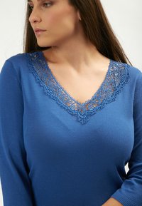 Maglione blu lavorato a maglia con scollo a V e dettagli in pizzo. Maniche lunghe con texture liscia e forma aderente.