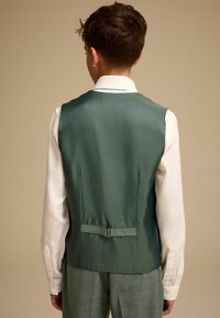 Grüne Satin-Weste mit glatter Textur, verstellbarem Riemen auf der Rückseite, getragen über einem weißen Hemd mit Kragen, kombiniert mit hellgrünen Hosen.