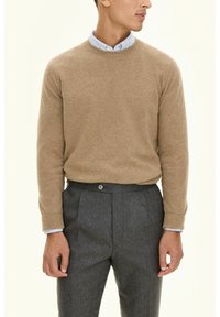 Beżowy sweter z dzianiny z prążkowanymi mankietami, noszony na jasnoniebieskiej koszuli z kołnierzykiem. Szare dopasowane spodnie z plisowanym przodem i detalem w postaci guzika w talii.