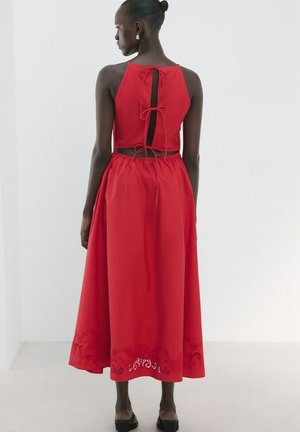 Donna che indossa un vestito rosso senza maniche con schiena scoperta legata da tre fiocchi, vita arricciata e orlo con ritagli floreali, in piedi contro un muro bianco.