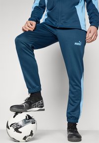 Námornícke športové nohavice s svetlomodrými bočnými pruhmi, elastickými manžetami a logom PUMA. Čierne športové topánky položené na čierno-bielom futbalovom míči.