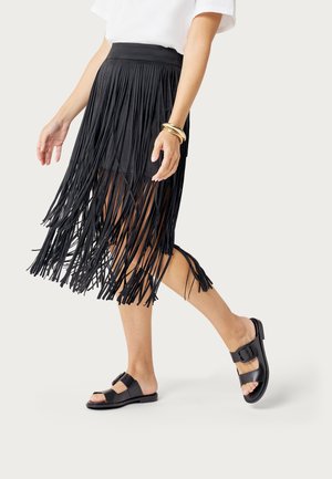 Donna che cammina indossando una gonna nera con frange, un top bianco, sandali neri e braccialetti dorati al polso destro.