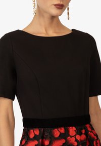 Schwarzes Kleid mit rundem Ausschnitt, kurzen Ärmeln und einem Samt-Taillengürtel, ergänzt durch ein rotes Blumenmuster auf dem Rock.