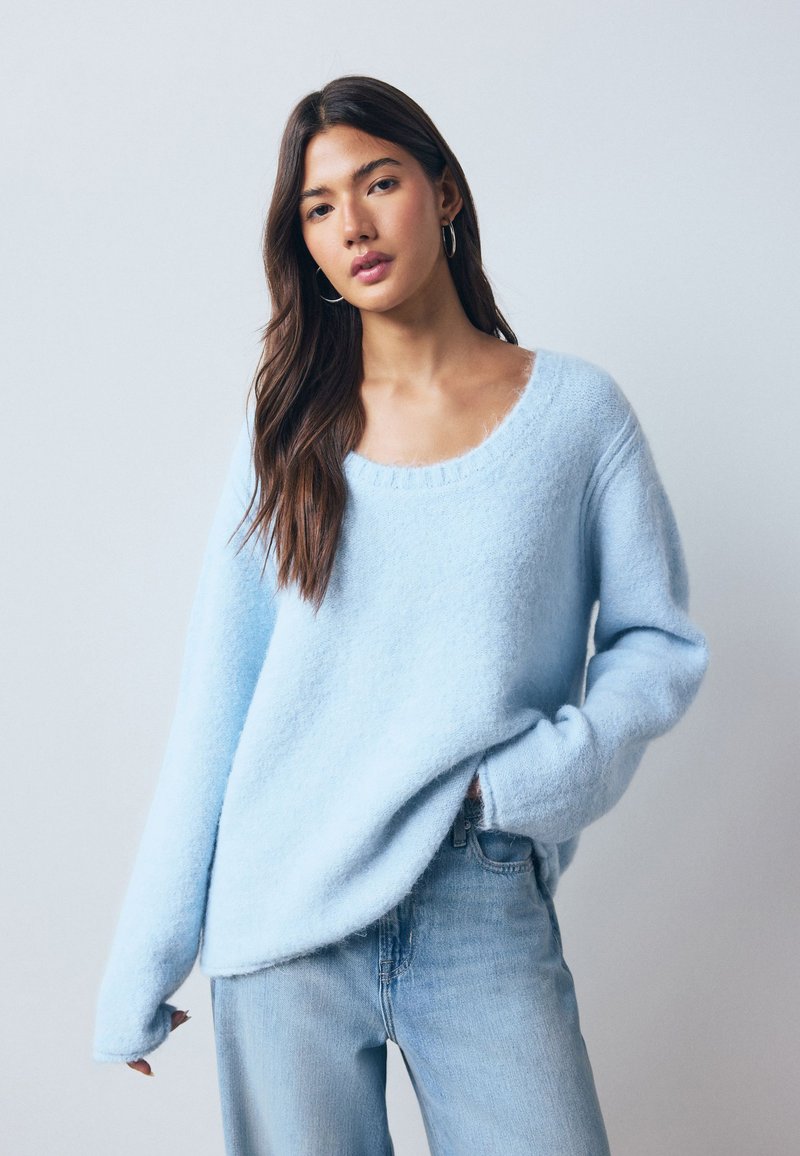Sky blue oversized sweater made of soft, textured material with a wide neckline and long sleeves, paired with light blue denim jeans.