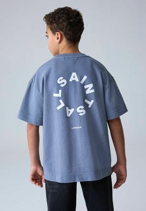 Boy with short curly hair wears loose blue t-shirt with white circular text "SAINT SAL" and "LONDON" standing against plain background.