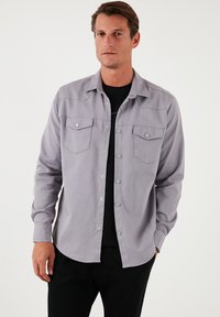 Cămașă gri, cu mânecă lungă, cu nasturi tip snap, guler ascuțit și două buzunare frontale. Textură moale, cu o culoare uniformă și un tiv curbat.