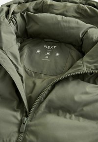 Jachetă puffer verde măsliniu, interior cu o textură netedă, având un fermoar complet, eticheta mărcii și simbolurile de îngrijire imprimate pe căptușeală.