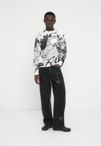 Μαύρο και λευκό γραφικό φούτερ με φαρδιά μαύρα τζιν παντελόνια διακοσμημένα με τρουκς. Το μοντέλο φορά μαύρα παπούτσια, στέκεται μπροστά από ένα λευκό φόντο.