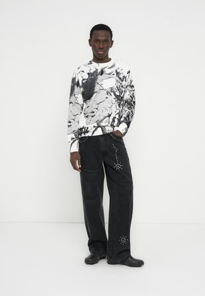 Crno-bijela grafička majica s dugim rukavima u kombinaciji s labavim crnim trapericama s ukrasnim zakovicama. Model nosi crne cipele i stoji pred bijelom pozadinom.