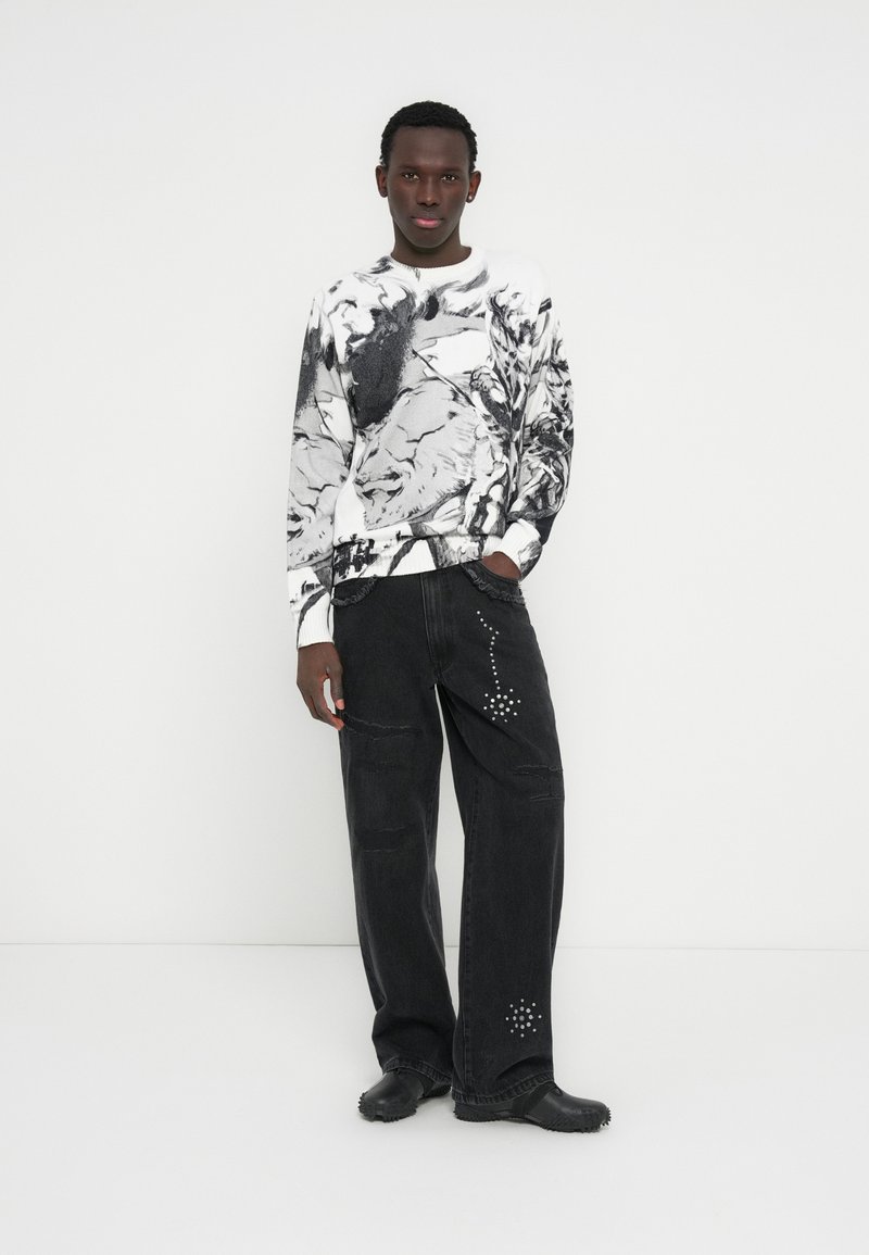 Μαύρο και λευκό γραφικό φούτερ με φαρδιά μαύρα τζιν παντελόνια διακοσμημένα με τρουκς. Το μοντέλο φορά μαύρα παπούτσια, στέκεται μπροστά από ένα λευκό φόντο.