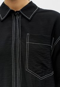 Chemise noire à boutons mettant en valeur des surpiqûres blanches en contraste sur le col, le devant, la poche et les coutures des manches, portée par une personne.