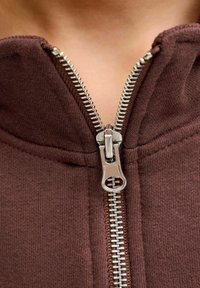 Haut à capuche zippé marron avec une fermeture éclair métallique, un tissu texturé et un col côtelé. La fermeture éclair est partiellement ouverte.