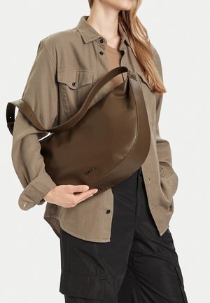 Žena v béžovém saku a černých kalhotách drží velkou hnědou koženou kabelku přes tělo.