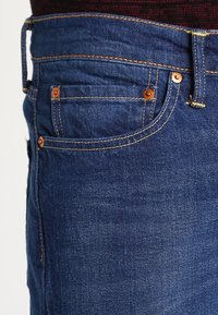 Jeans de mezclilla azul oscuro con costuras naranjas y herrajes en tono cobre. Incluyen un bolsillo delantero con detalles de costura visibles.