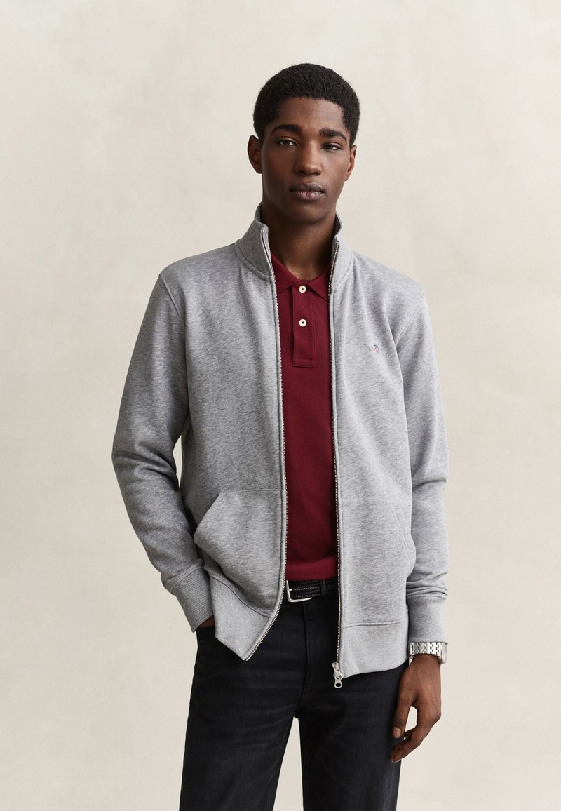 Jeune homme portant une veste zippée grise par-dessus un polo bordeaux, avec un pantalon noir et une montre argentée, debout devant un fond uni.