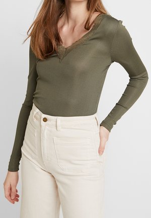 Frau trägt ein olivgrünes, geripptes Langarmshirt mit Spitzeneinsatz am V-Ausschnitt und hochtaillierte, cremeweiße Hose mit Vordertaschen.