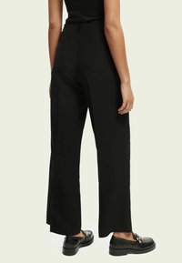 Hoge zwarte broek met een wijde pijpdesign, voorzien van twee achterzakken en gedragen met zwarte instapschoenen. Glad, effen materiaal.