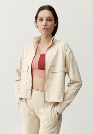 Donna che indossa giacca beige con zip e pantaloni, con reggiseno sportivo rosa e rosso, in piedi con le mani nelle tasche su sfondo neutro.
