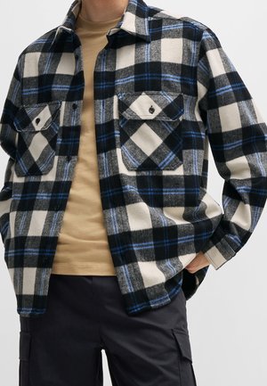 Homme portant une chemise à carreaux noire, blanche et bleue à boutons par-dessus un t-shirt beige et un pantalon cargo noir, debout avec une main dans la poche.