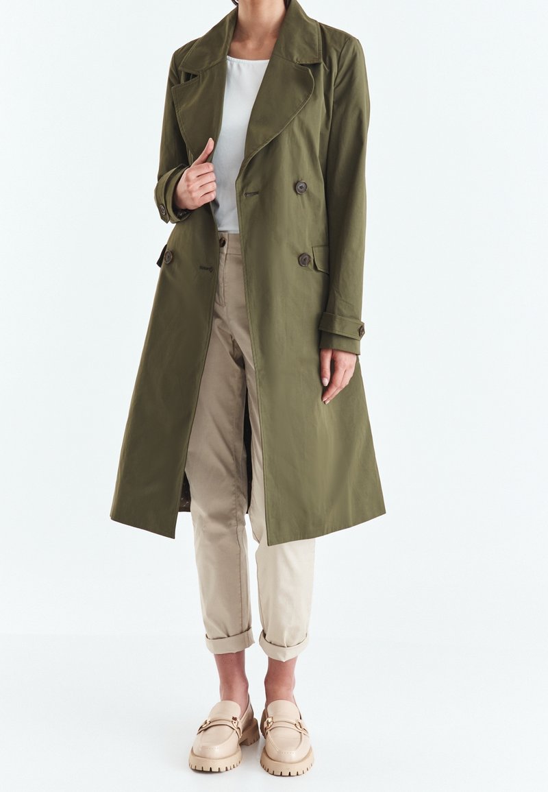 Trench-coat vert olive à double boutonnage, grands revers et boutons. Associé à un pantalon court crème et des mocassins beige.