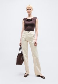 Γυναίκα με κοντά ξανθά μαλλιά, φοράει αμάνικο καφέ τοπ, κρεμ παντελόνι φαρδύ, καφέ παπούτσια και κρατά μια μεγάλη καφέ τσάντα ώμου.