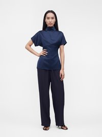 Femme debout avec une main sur la hanche, portant un haut bleu marine à col montant et manches courtes, un pantalon large et sombre, et des sandales noires, sur un fond uni.