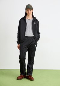 Fato desportivo preto com casaco com fecho e calças com cintura elástica. T-shirt cinza por baixo. Sapatos castanhos e boné verde com logótipo.