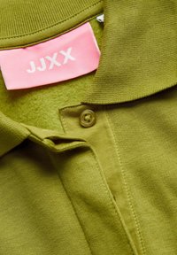Olive green polo shirt with a textured fabric, folded collar, and a pink tag labeled "JJXX." Features a single button closure.