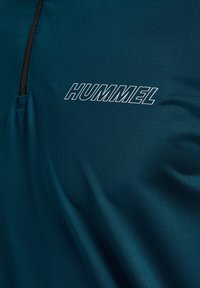 Mørk teal sportsstof med sort lynlås og hvid omridset tekst "HUMMEL" på brystområdet.