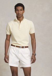 Polo em amarelo pálido com mangas curtas, combinado com calções brancos. Um cinto de couro castanho e um relógio de pulso são acessórios notáveis.