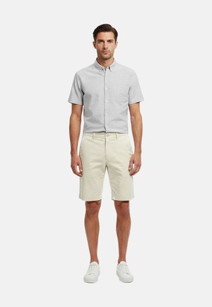 Hombre con camisa gris claro de manga corta abotonada, pantalones cortos beige hasta la rodilla y zapatillas blancas, de pie sobre fondo blanco.