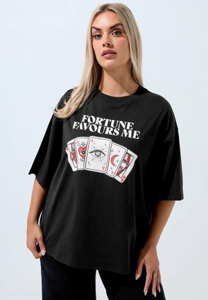Lyshåret kvinde iført en løs sort t-shirt med teksten "FORTUNE FAVOURS ME" og illustrerede spillekort med mystiske symboler.