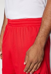 Shorts de sport en filet rouge avec une ceinture élastique, présentant un motif texturé et une coupe décontractée. Main posée sur les shorts.
