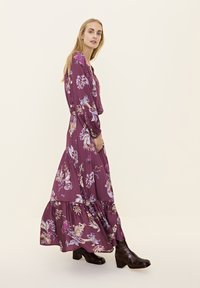 Lila Blumen-Maxikleid mit langen Ärmeln, gestuftem Rock und geraffter Taille. Getragen mit dunkelbraunen Stiefeletten auf einem neutralen Hintergrund.