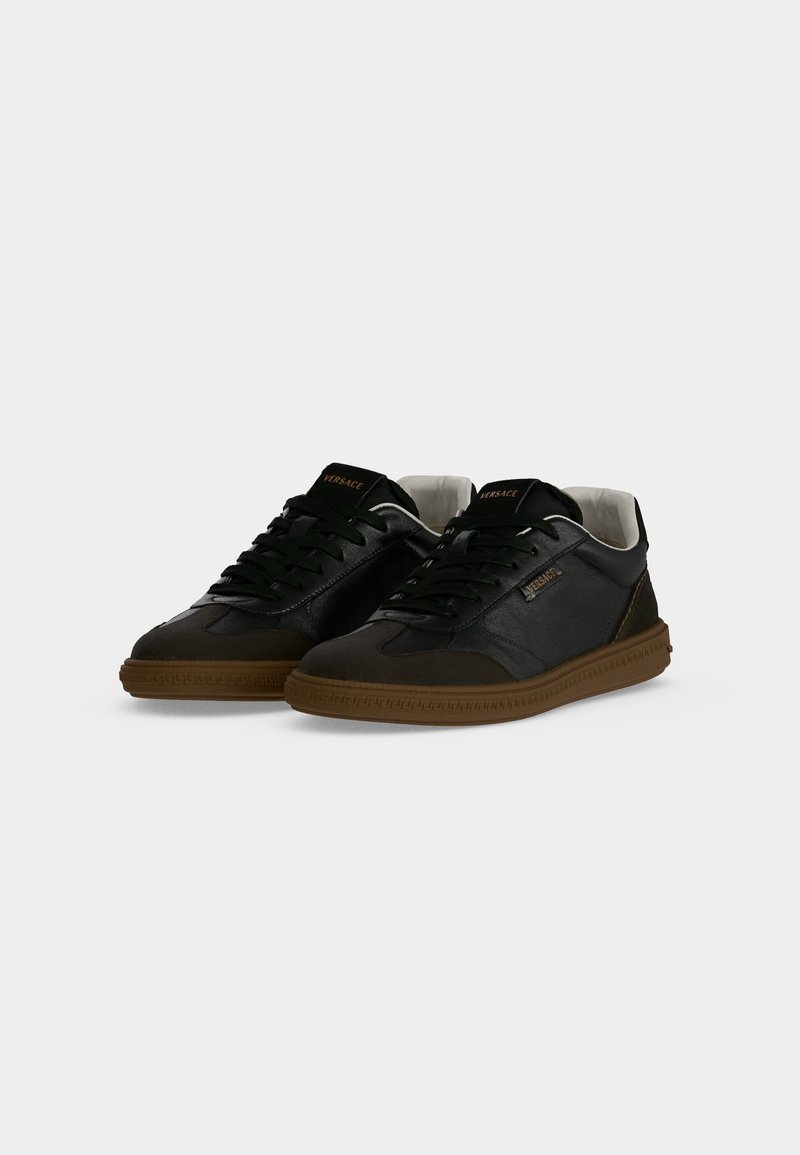 Coppia di sneakers nere in pelle basse con suole marroni, lacci neri e logo Versace sul lato e sulla linguetta su sfondo bianco.
