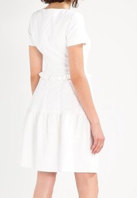 Vestido blanco de manga corta con un corsé ajustado, cintura con volantes y falda acampanada. La tela texturizada y las líneas limpias crean un estilo minimalista.