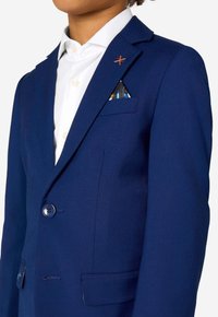 Zils bleizeris ar izgrieztām atlokām, vienu pogu un dekoratīvu oranžu krustšuvīti. Satur rakstainu kabatas lakatiņu krūšu kabatā.