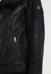 Czarna kurtka z sztucznej skóry o teksturowanym wykończeniu, z boczną kieszenią na zamek oraz gładkim wykończeniem z tkaniny wzdłuż krawędzi.