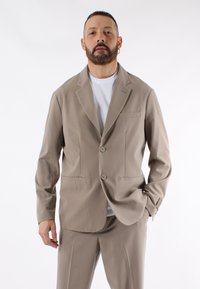 Completo beige chiaro con due bottoni e colletto a scollo. Presenta una tasca sul petto e due tasche frontali. Indossato su una camicia bianca.