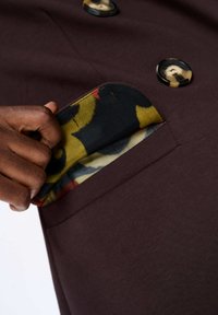 Bruine stoffen jaszak onthuld, gevoerd met een gemusterde stof met gele, rode en marineblauwe wervelingen. Zichtbare ronde zwarte knopen.