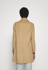 Manteau en laine beige avec col cranté, coupe ajustée, manches longues et fente à l'arrière. Texture lisse, longueur jusqu'aux genoux, design minimaliste.