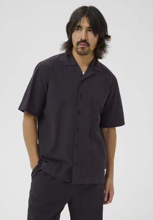 Homme aux cheveux bruns mi-longs et moustache, portant une chemise à manches courtes foncée boutonnée et un pantalon assorti, devant un fond uni.