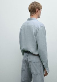 Chemise à manches longues gris clair avec col, poignets boutonnés et empiècement au dos, associée à un jean en denim gris clair avec deux poches arrière.