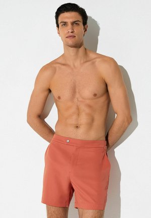 Tumejuukseline mehevarv seina ees, valge taustaga, seljas korallvärvi ujumispüksid, käed selja taga.