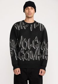 Pull en tricot noir avec de grands motifs en texte gris. Col rond, poignets et ourlet côtelés. Coupe simple et décontractée, porté avec un pantalon noir.