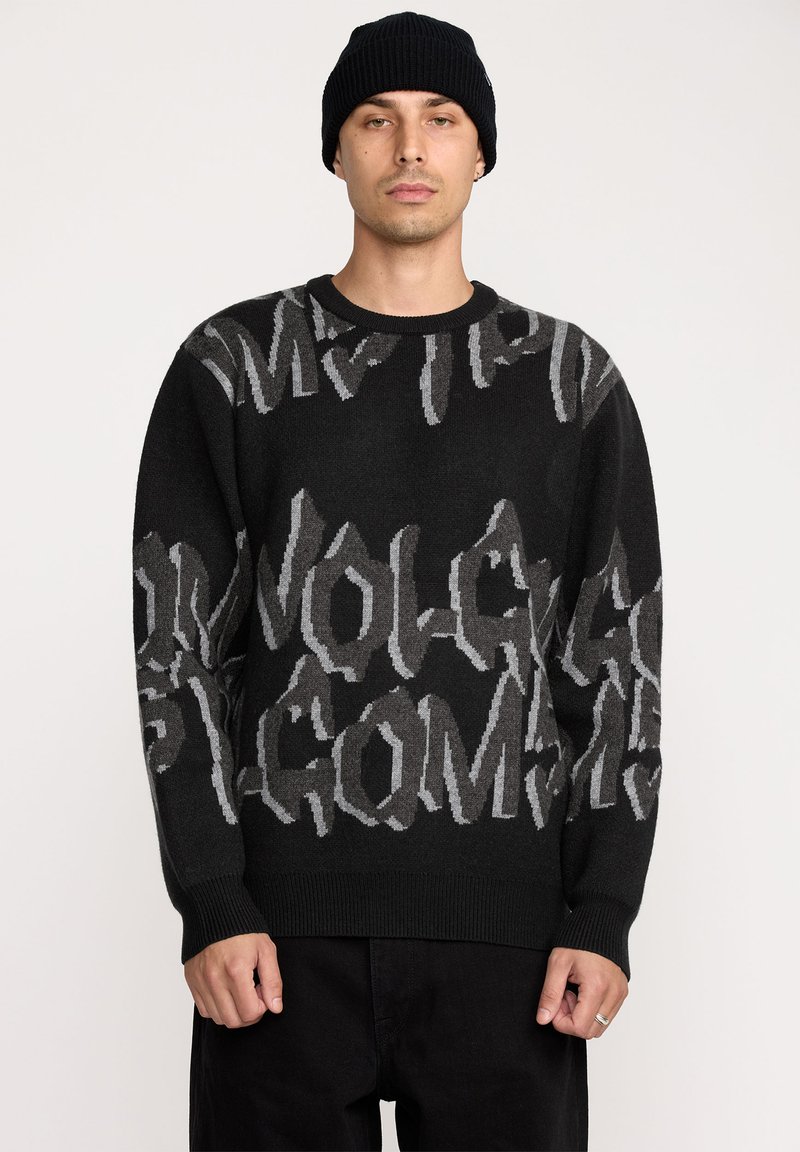 Pull en tricot noir avec de grands motifs en texte gris. Col rond, poignets et ourlet côtelés. Coupe simple et décontractée, porté avec un pantalon noir.