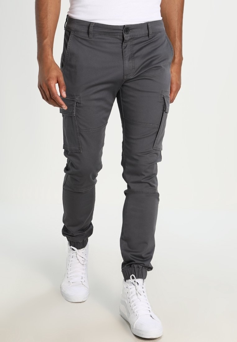 Homme portant un pantalon cargo gris foncé avec des poches latérales et des poignets élastiques, associé à des baskets blanches montantes, debout devant un fond uni.