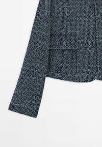 Giacca blu scuro con tessuto strutturato e intrecciato. Presenta una tasca sul petto e maniche dal taglio dritto.